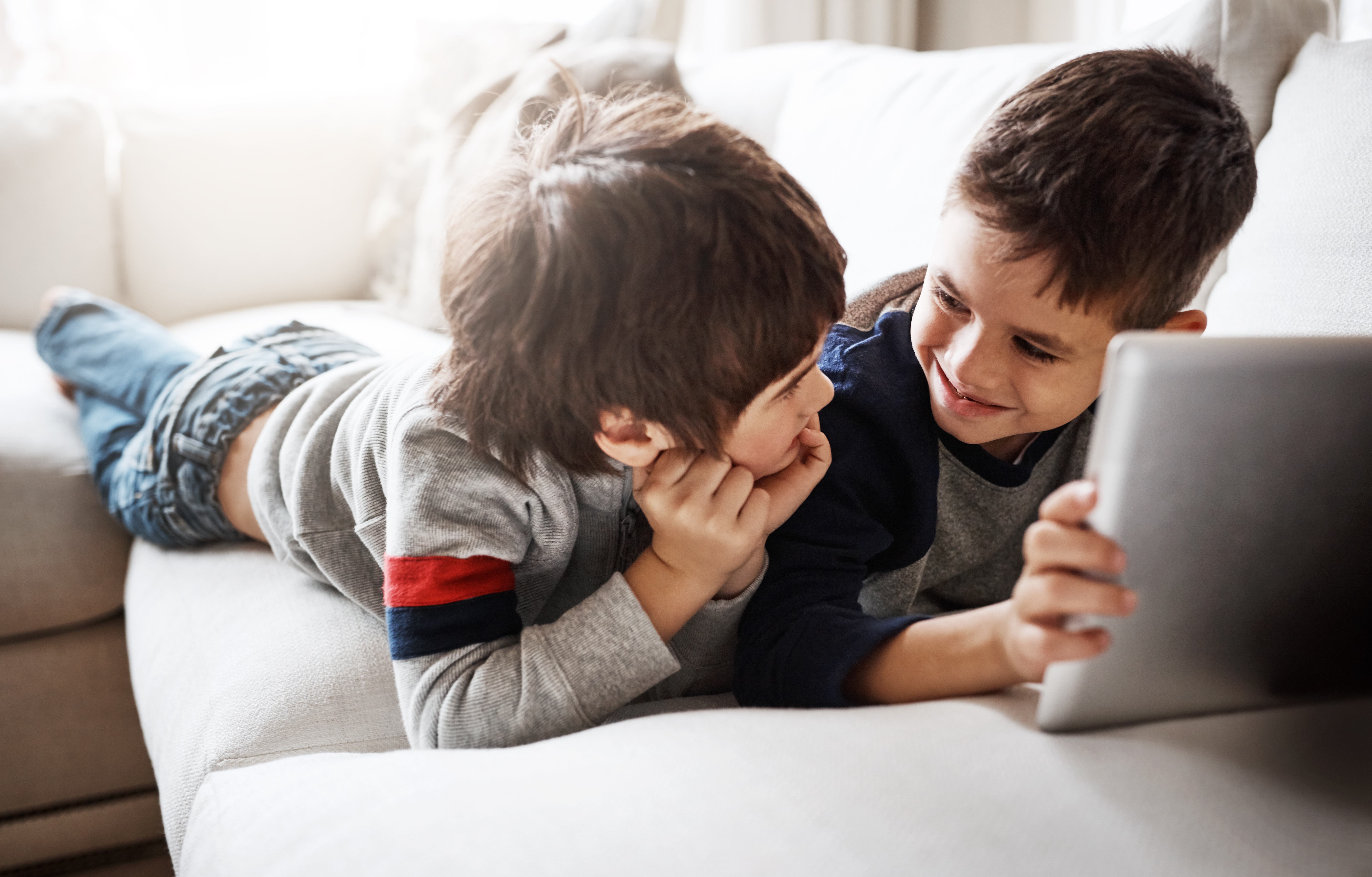 Zwei junge Kinder liegen nebeneinander auf dem Sofa auf dem Bauch liegen und nutzen gemeinsam ein Tablet. Sie lachen sich an.