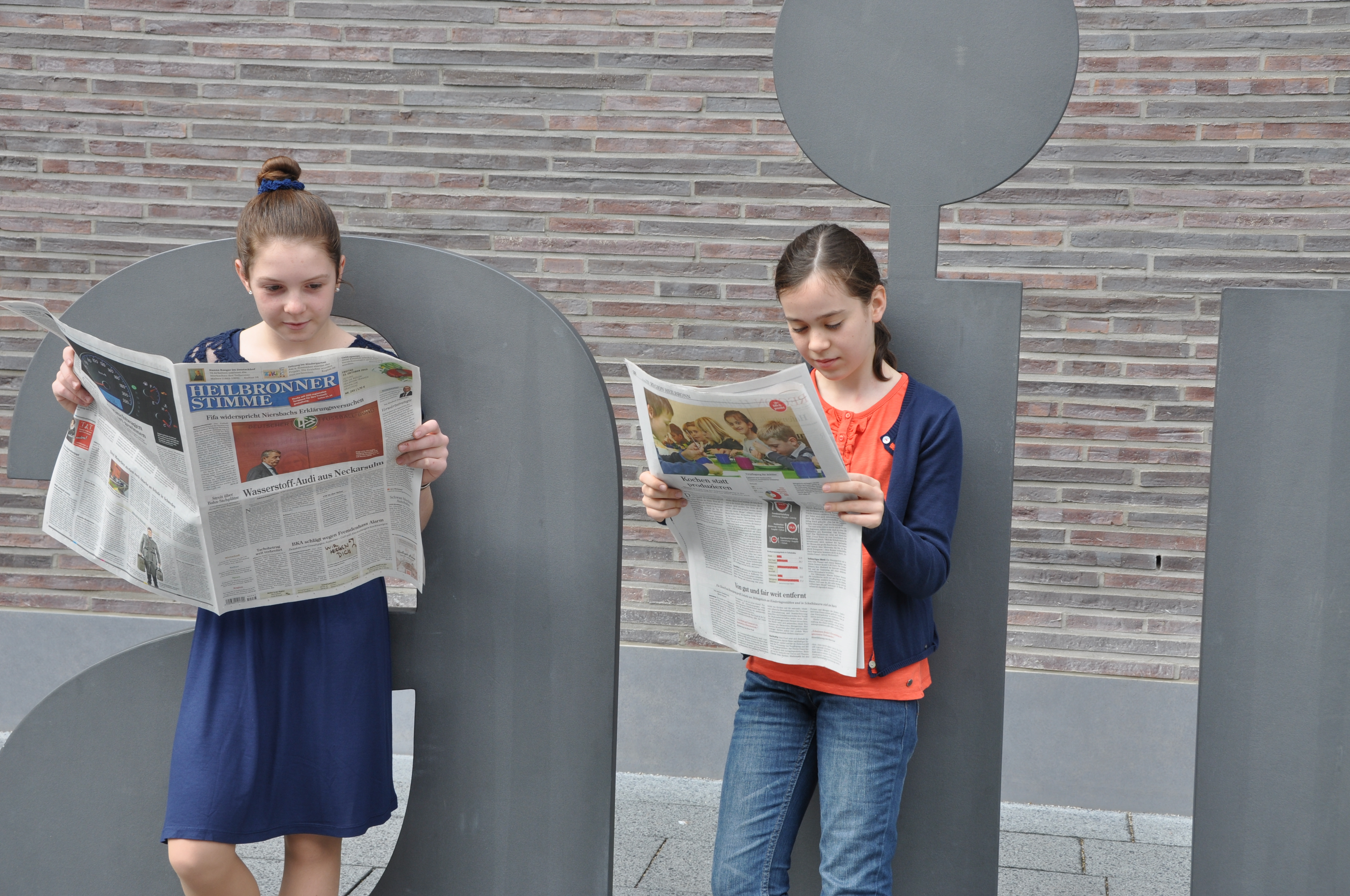 Zwei junge Schülerinnen stehen vor dem aim Buchstaben vor dem aim-Gebäude und lesen die Heilbronner Stimme.