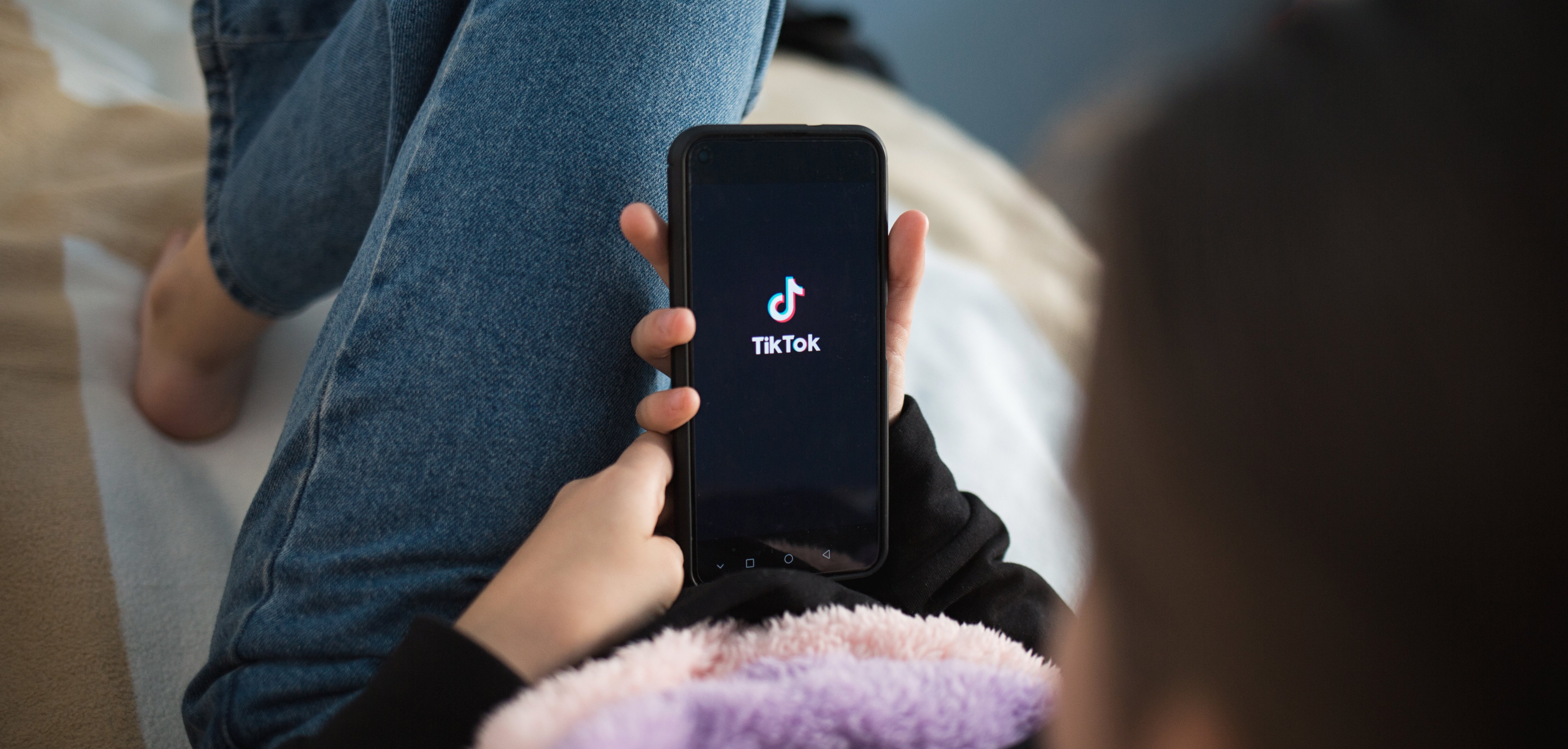 Junges Kind liegt auf dem Bett und hält ein Handy in der Hand. Das Logo von der Social Media App TikTok ist auf dem Bildschirm.
