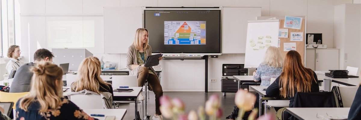 Dozentin sitzt mit dem Tablet in der Hand auf dem Tisch in einem Raum voller Teilnehmerinnen und Teilnehmer. Das Smartboard im Hintergrund zeigt eine Grafik.